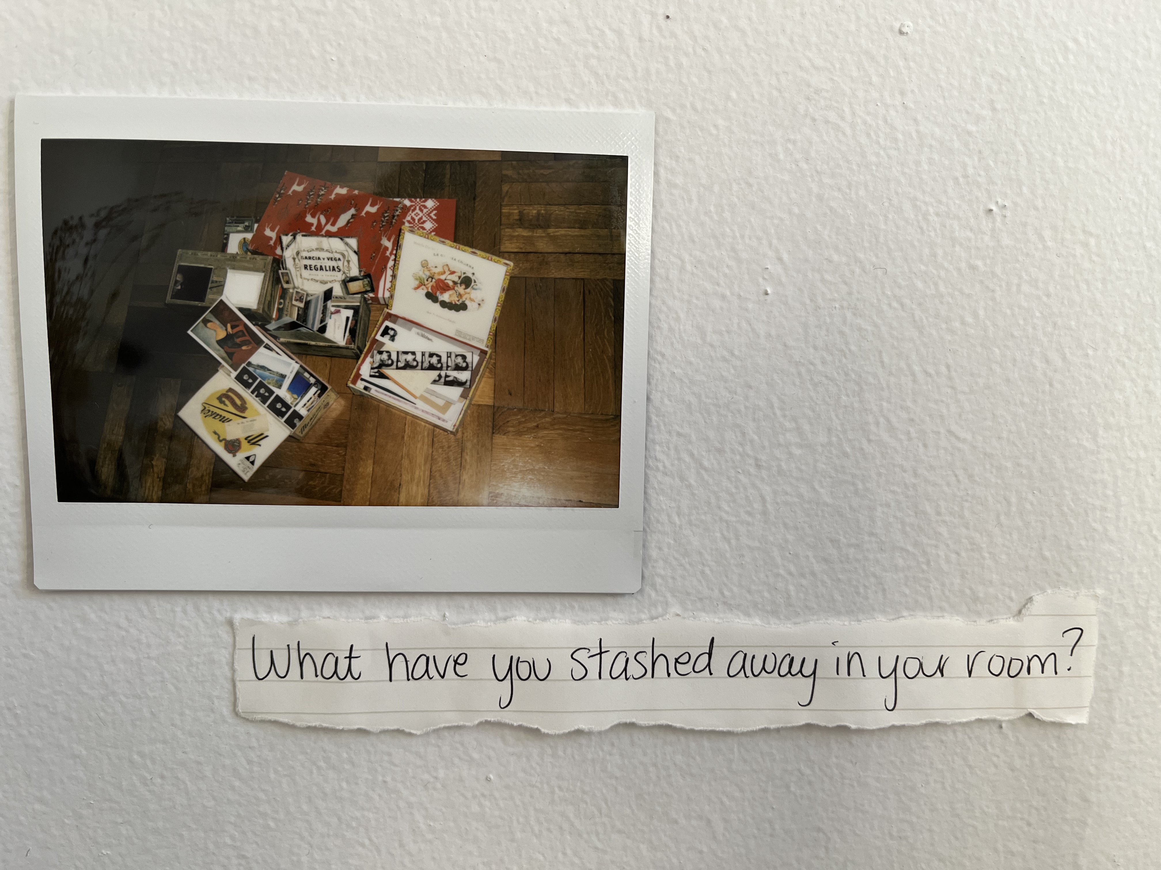 A hand-written note on the wall reads, What have you stashed away in your room?

A polaroid above the note shows the boxes in the previous polaroid, now open so we can see some of their contents. They are filled with photo strips, polaroids, and postcards. 