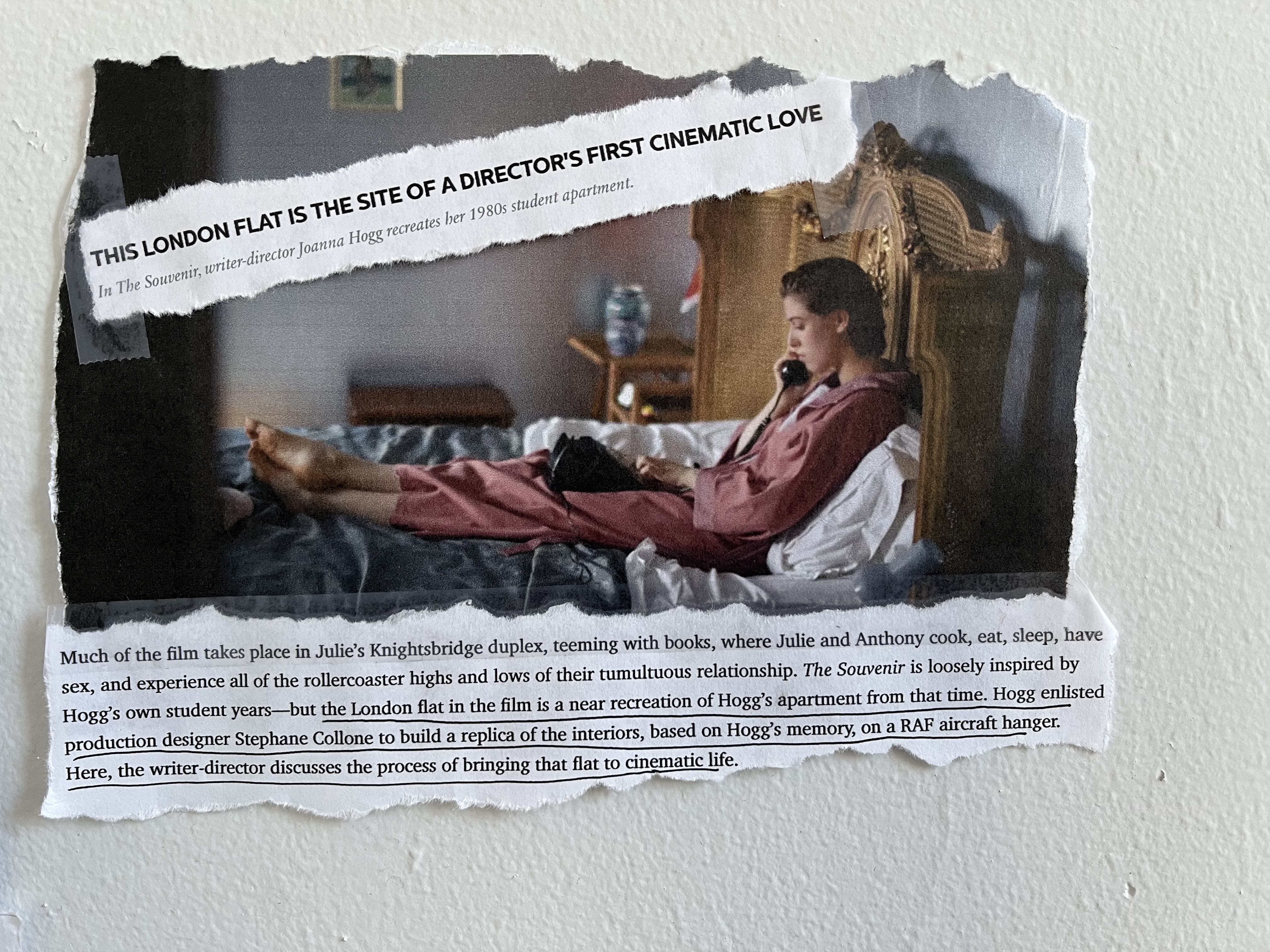 Cutouts from a magazine article are taped to wall. A photo shows a still from the film The Souvenir, of the young woman protagonist sitting on her bed on the telephone. The text reads:

This London Flat is the Site of a Director's First Cinematic Love
In The Souvenir, writer-director Joanna Hogg recreates her 1980s student apartment.

Much of the film takes place in Julie's Knightsbridge duplex, teeming with books, where Julie and Anthony cook, eat, sleep, have sex, and experience all of the rollercoaster highs and lows of their tumultuous relationship. The Souvenir is loosely inspired by Hogg's own student years — but the London flat in the film is a near recreation of Hogg's apartment from that time. Hogg enlisted production designer stephane Collone to build a replica of the interiors, based on Hogg's memory, on a RAF aircraft hanger. Here, the writer-director discusses the process of bringing that flat to cinematic life. 