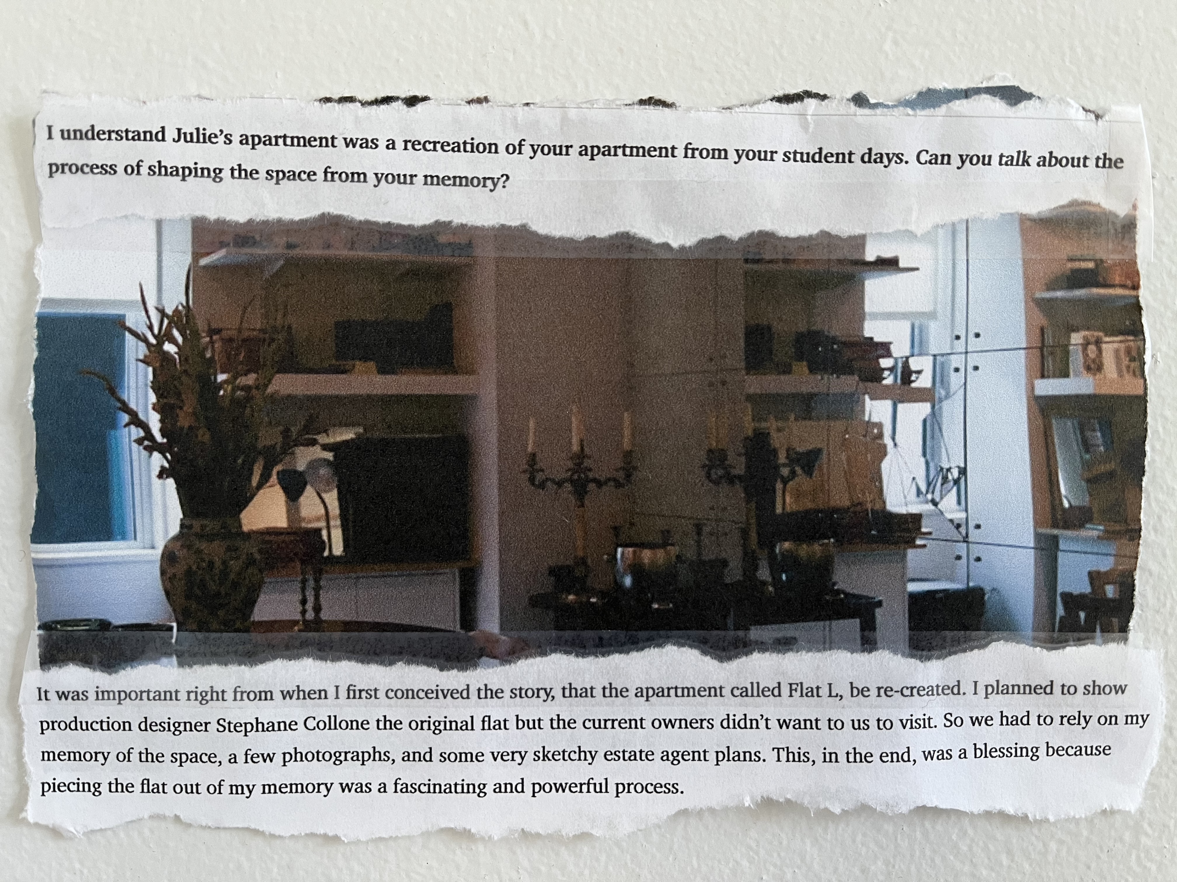 Another section of the magazine article about The Souvenir is taped to the wall. An image shows a corner of a room, furnished with tables, shelves, and objects including a candlestick and vase. The text, part of an interview with Joanna Hogg, reads:

Interviewer: I understand Julie's apartment was a recreation of your apartment from your student days. Can you talk about the process of shaping the space from your memory?

Hogg: It was important right from when I first conceived the story, that the apartment called Flat L, be re-created. I planned to show production designer Stephane Collone the original flat but the current owners didn't want us to visit. So we had to rely on my memory of the space, a few photographs, and some very sketchy estate agent plans. This, in the end, was a blessing because piecing the flat out of my memory was a fascinating and powerful process. 