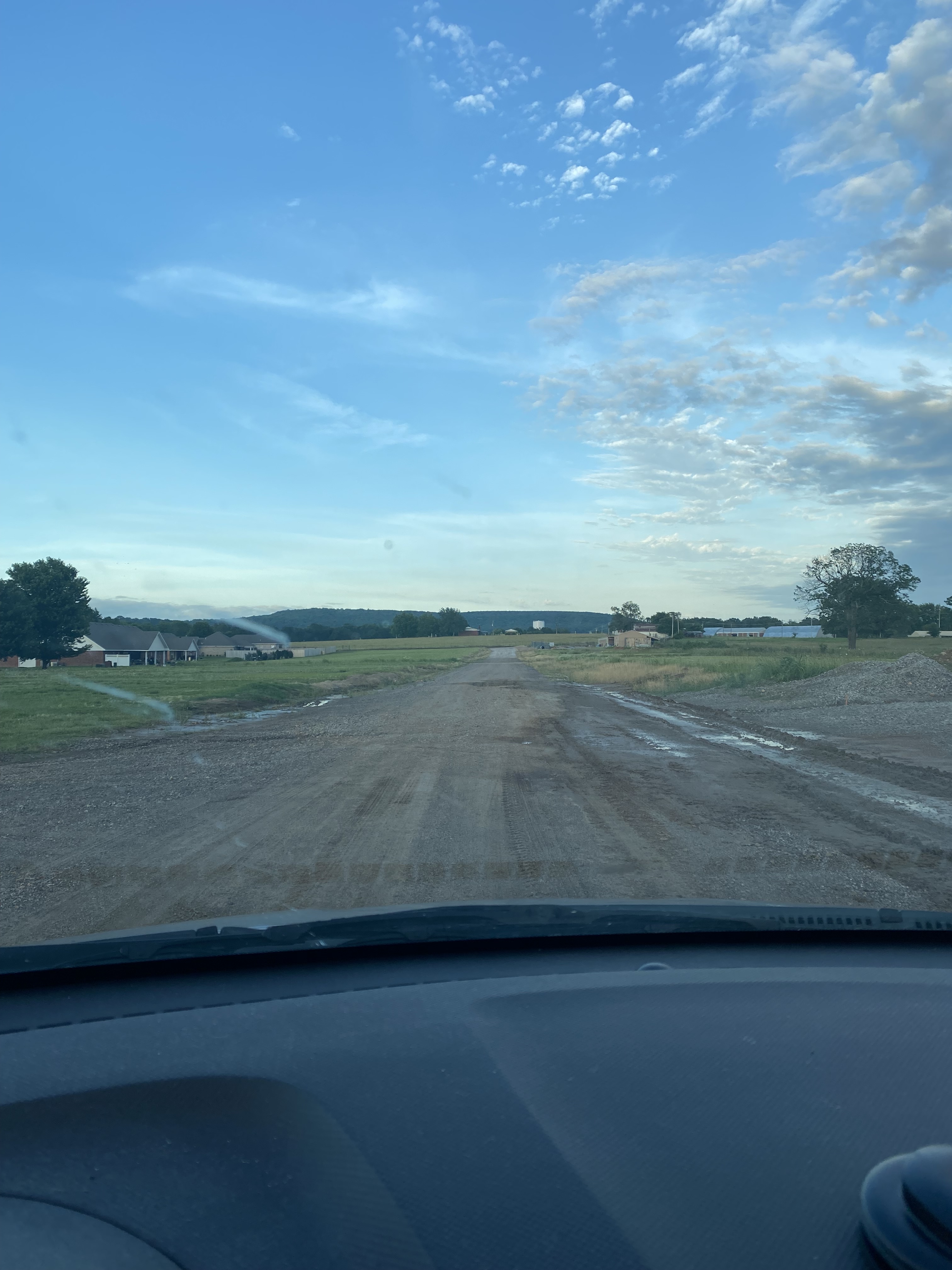 A photo taken from the front seat of a car. Beyond the dashboard, through the windshield, a long dirt road extends into the distance, surrounded by fields, with a couple houses here and there. Big blue sky with a few tray clouds.