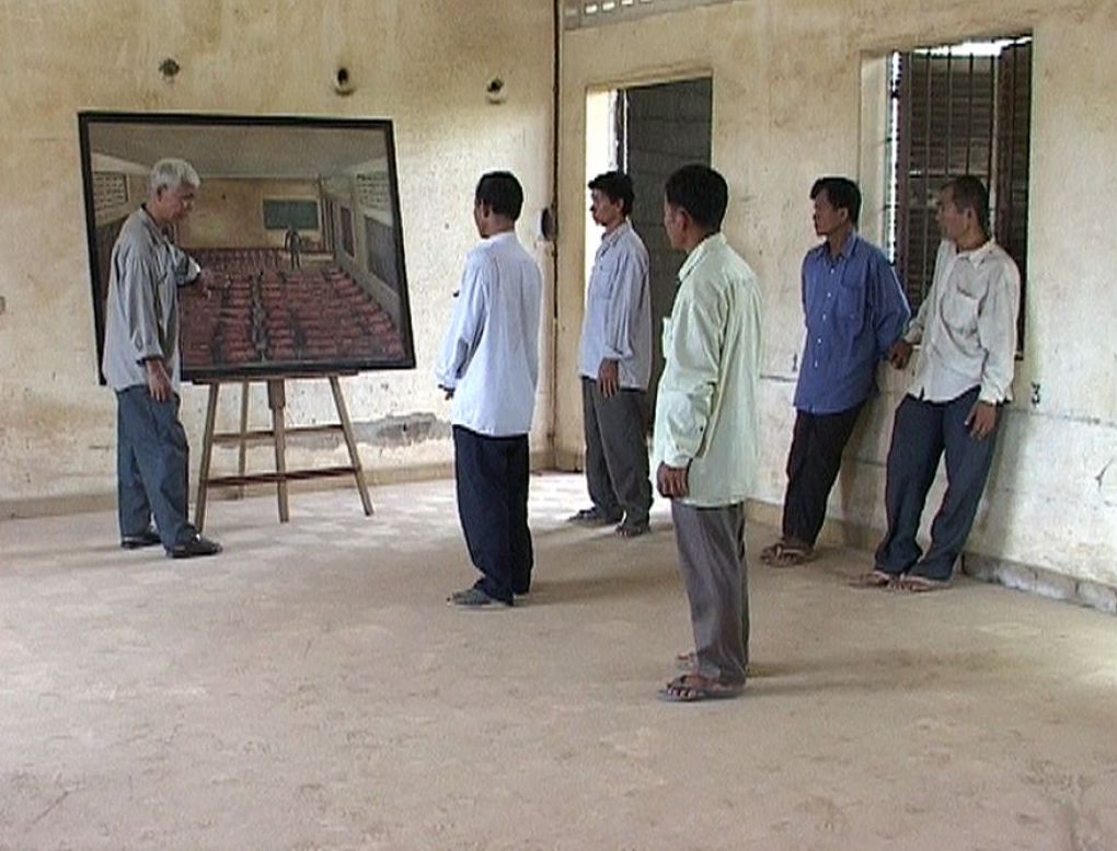 In a large, bare room, a group of five men look on while one man gestures towards a painting on an easel. 