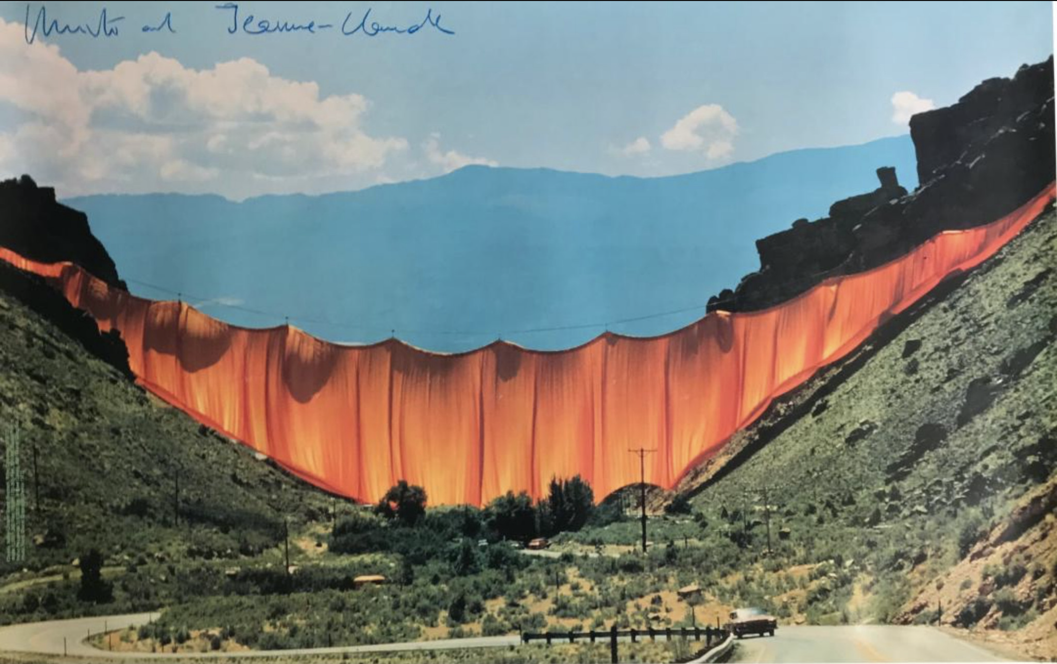 A gigantic orange curtain spans a green valley in front of a blue mountain  with a highway running through. 